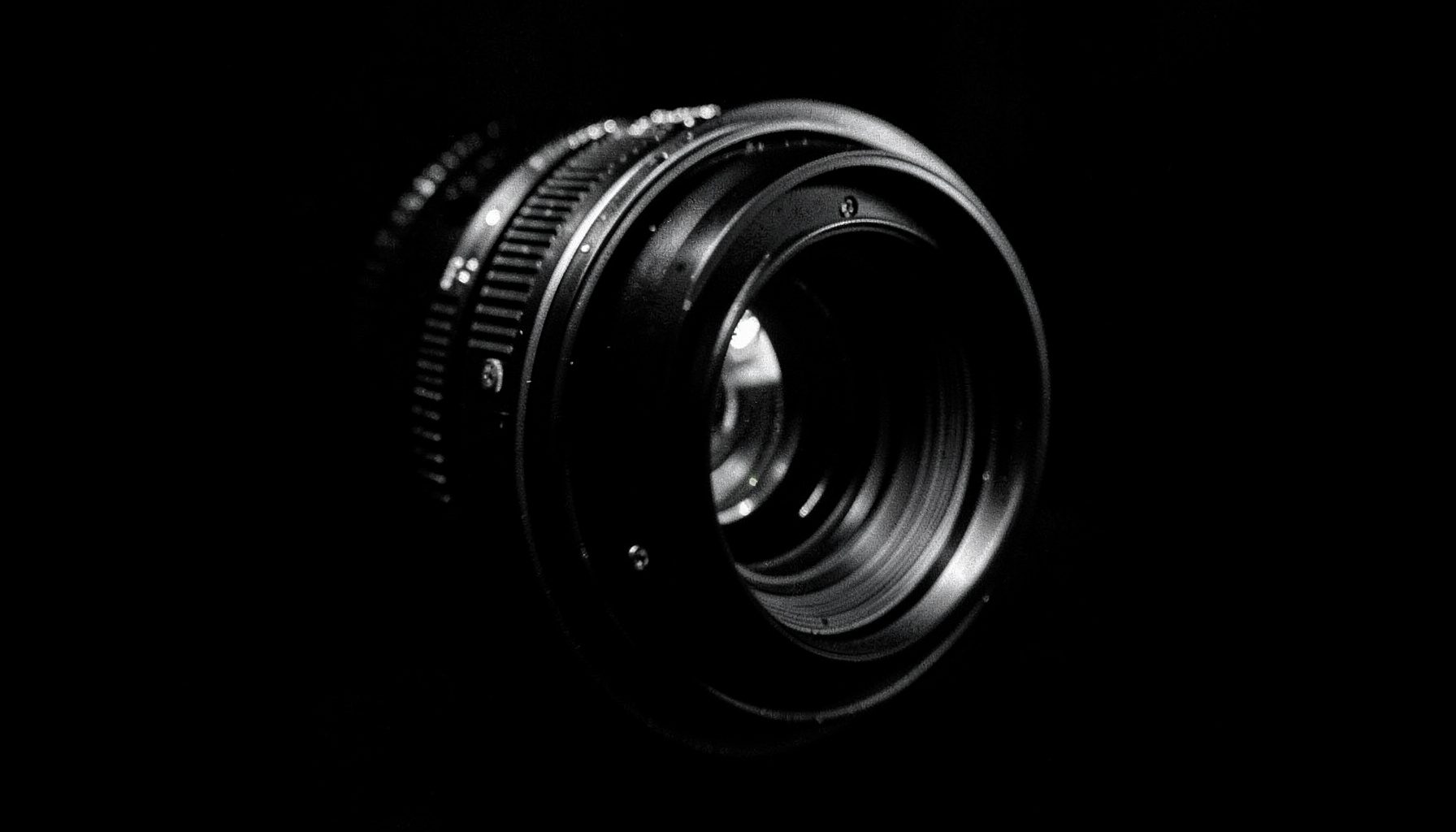 A dark, atmospheric duotone photograph of a camera lens seen from an angle, with deep charcoal tones and subtle silver highlights against a black background