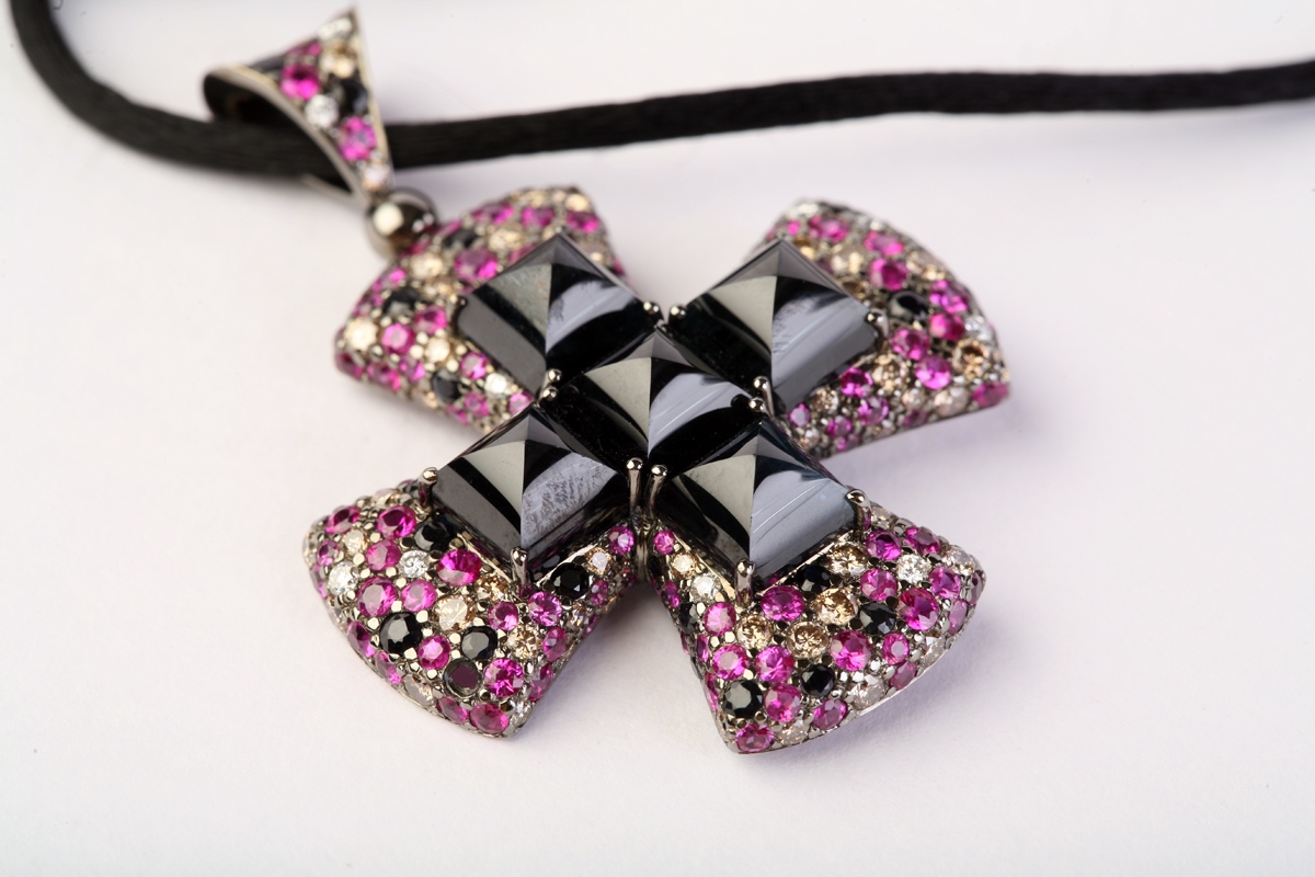 Close-up of an ornate cross-shaped pendant covered in pink and clear gemstones with black faceted stones, on a white background